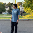 Person wearing a blue t-shirt with text, standing on a street with trees and houses in the background.