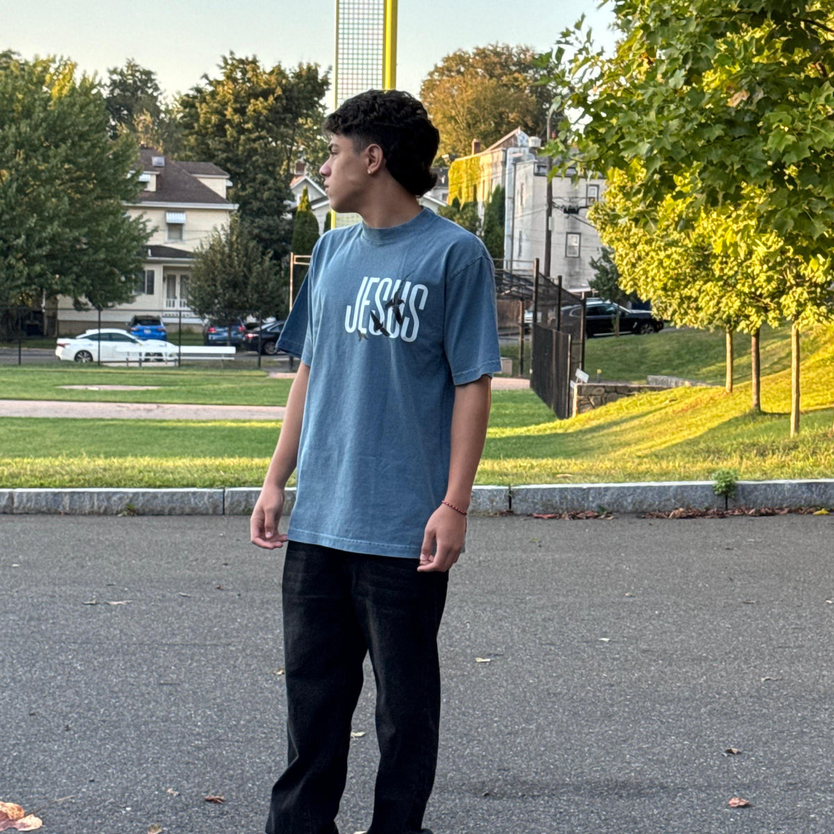 Person wearing a blue t-shirt with text, standing on a street with trees and houses in the background.
