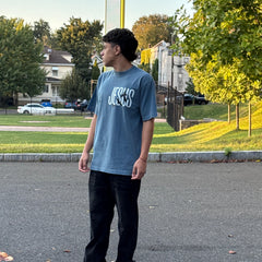 Person wearing a blue t-shirt with text, standing on a street with trees and houses in the background.