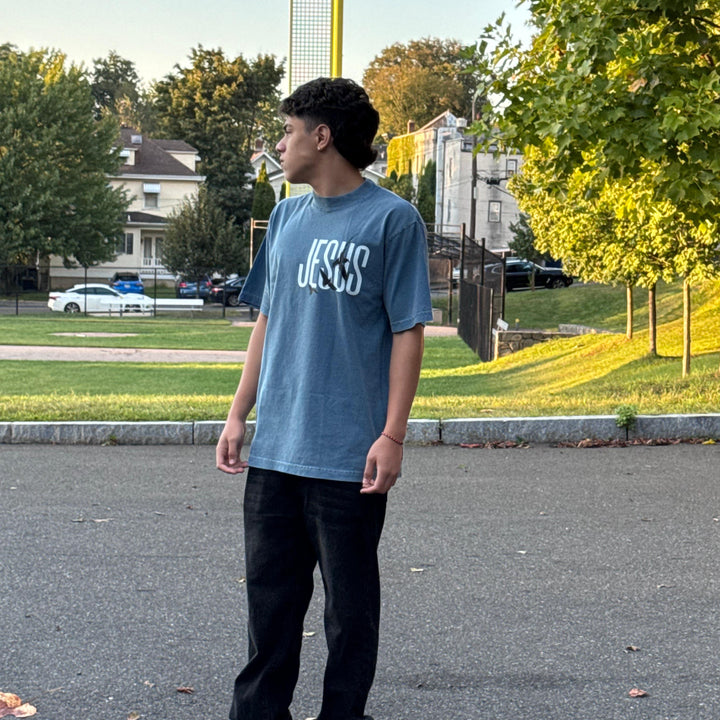 Person wearing a blue t-shirt with text, standing on a street with trees and houses in the background.