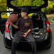 Person sitting in the open trunk of a car holding a basketball, with trees and road in the background.